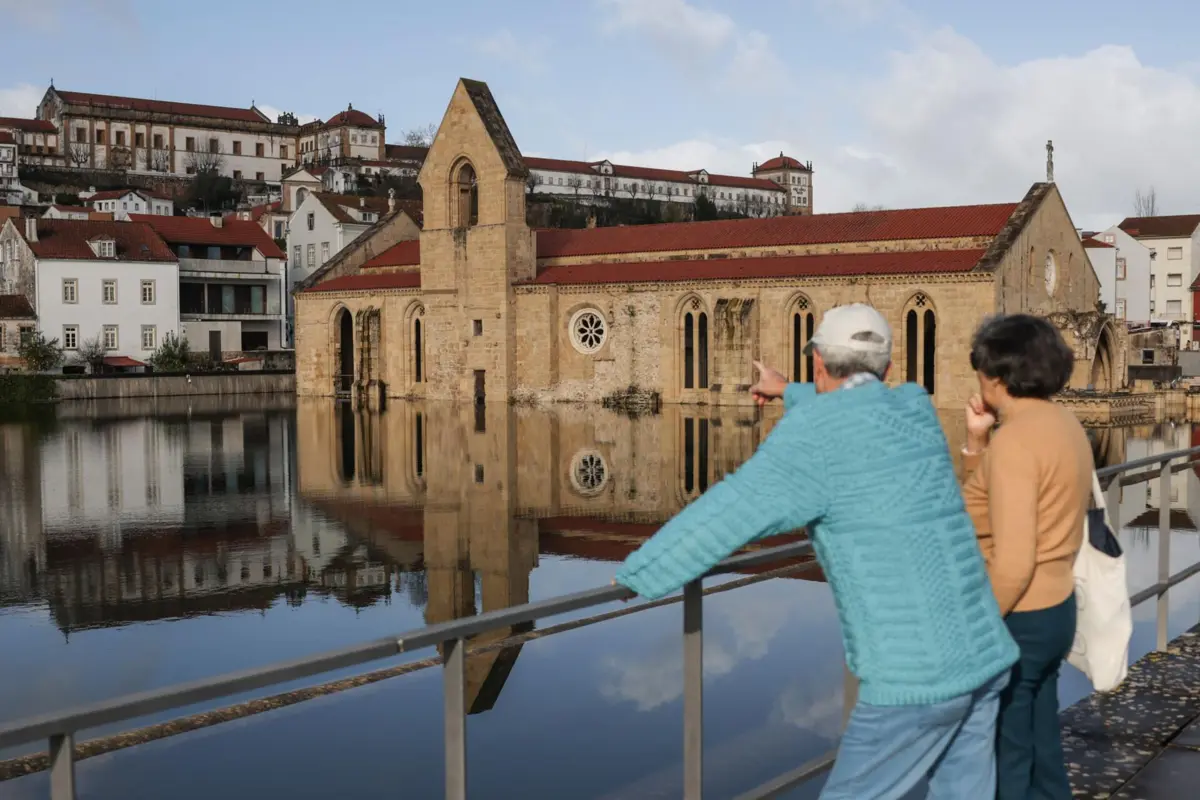 Populares observam o Mosteiro de Santa Clara-a-Velha inundado. A zona ribeirinha da margem esquerda esteve parcialmente inundada: o Mondego extravasou desde o Parque da Canção até às traseiras do Mosteiro de Santa Clara-a-Velha. em Coimbra, 14 de fevereiro de 2026. Na Ponte-Açude, o caudal do Mondego atingiu um máximo de 2.105 m3/s às 16:00 de quarta-feira - sensivelmente duas horas antes de ruir o dique da margem direita do Mondego que levou, depois, à queda de parte do tabuleiro da autoestrada 1 (A1) -, esteve em redor dos 2.000 m3 na madrugada de quinta-feira e tem vindo a baixar consistentemente, situando-se, pelas 10:00, nos 1.646. MIGUEL A. LOPES/LUSA